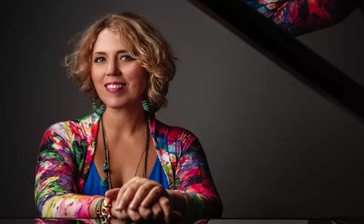 Portrait of pianist Gabriela Montero, smiling and seated at a piano, wearing a colorful floral cardigan.
