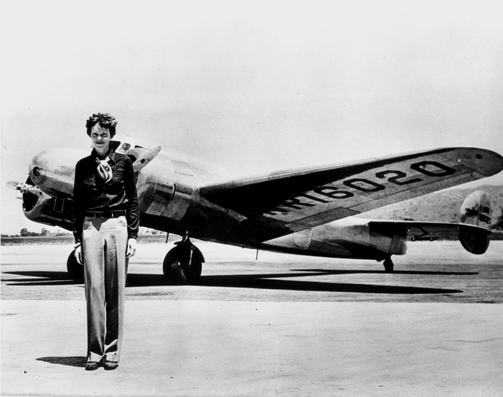 A woman standing in front of a vintage airplane on a runway, wearing a black blouse and light-colored trousers.