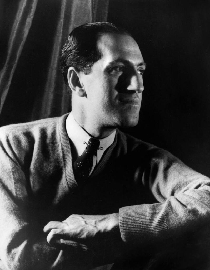 Black and white portrait of a man in a sweater, posed thoughtfully with his hand resting on his arm, lit with dramatic shadows.