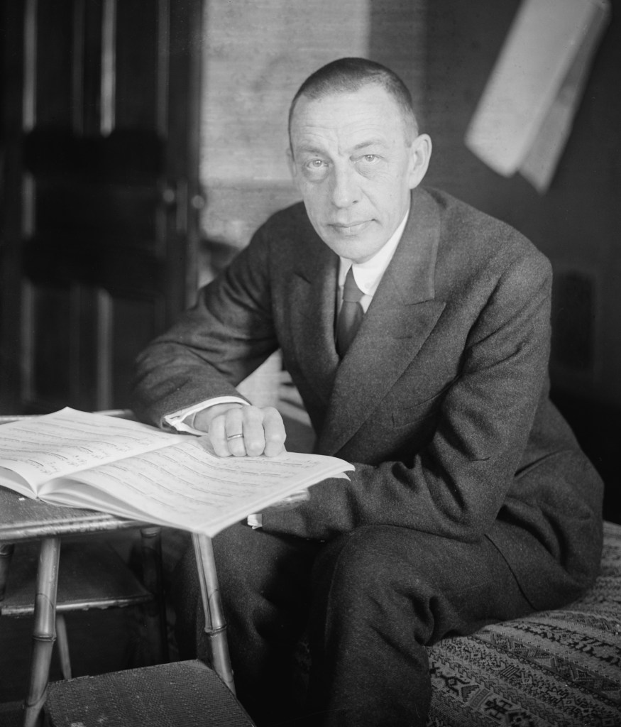 Black and white portrait of a man in a suit sitting at a table with music sheets, looking directly at the camera.