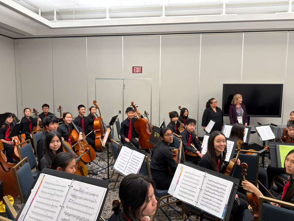 A group of young musicians seated in an orchestra setting, smiling and preparing for a performance. They are dressed in black attire with red ties, surrounded by music stands with sheet music.