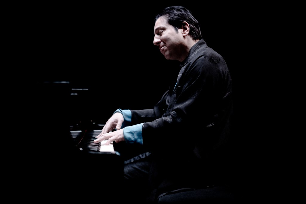 A pianist in a dark setting, playing a grand piano with focused expression, wearing a black outfit with blue accents.