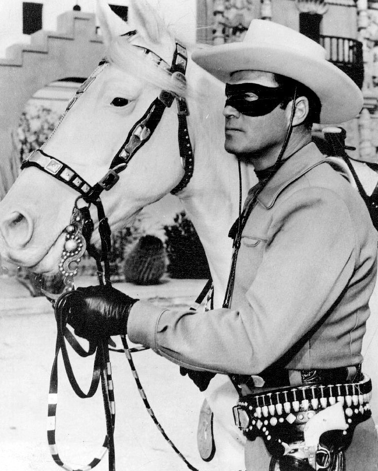 A black and white image of a masked cowboy in a light-colored shirt and wide-brimmed hat, holding the reins of a white horse. The cowboy has leather gloves and is adorned with a belt featuring various holsters and accessories.