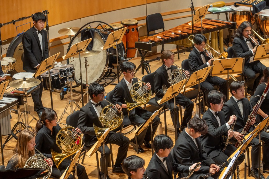 A group of young musicians wearing formal black attire is seated in an orchestra setting, playing various brass and woodwind instruments, with sheet music on stands in front of them. In the background, there are additional percussion instruments.