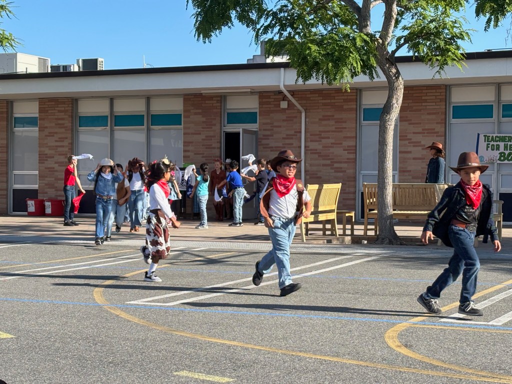 Group of children dressed in cowboy outfits participating in an outdoor activity at a school, with some wearing hats and bandanas.