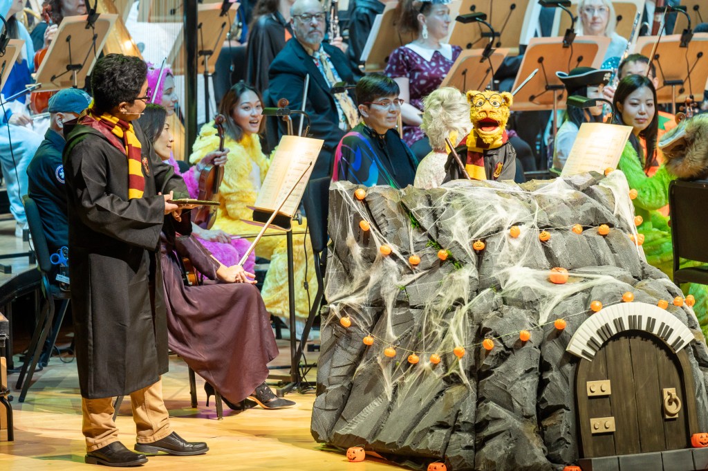 A performer in a Harry Potter costume stands beside a large, decorated rock stage with puppet characters and Halloween decorations, while musicians in the background, dressed in colorful costumes, play their instruments.
