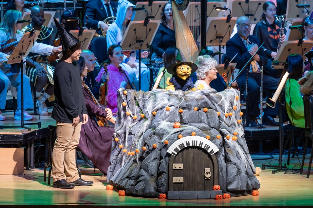 A performer in a black shirt and witch hat stands next to a rock-themed stage prop decorated with pumpkins and spider webs, featuring a puppet dressed as a witch. In the background, musicians in various costumes play orchestral instruments.