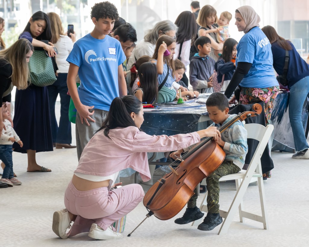 A young girl kneeling next to a boy playing a cello at a community event, with several children and adults engaging in various activities at tables in the background.