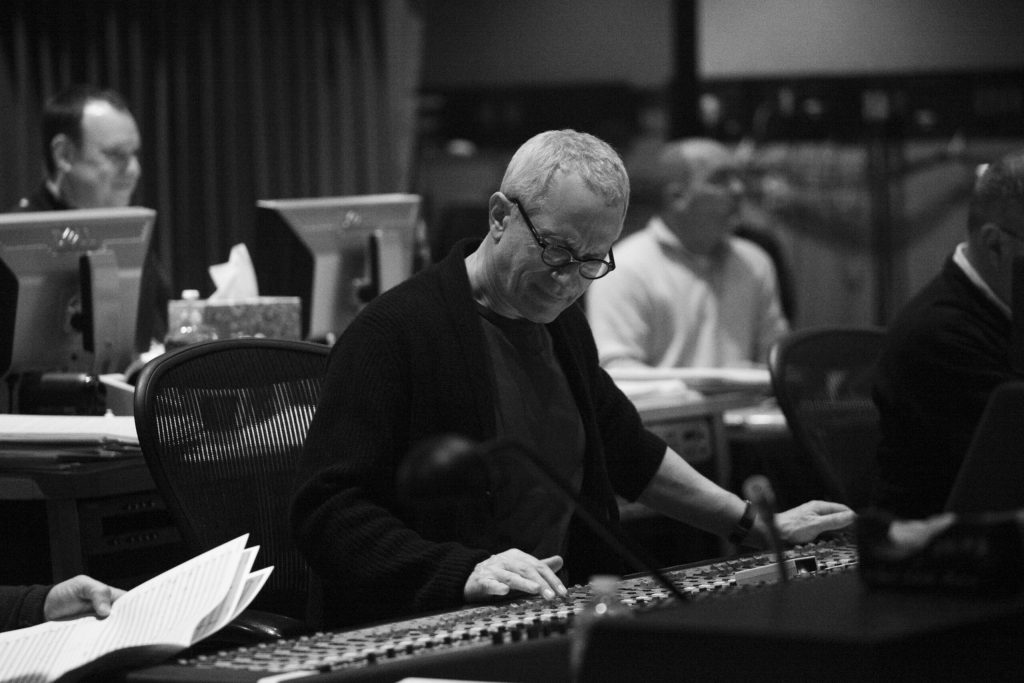 A black and white image of a music producer or sound engineer working at a mixing console, with several monitors and colleagues in the background.