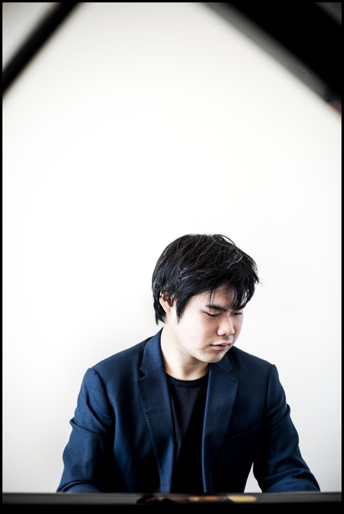 A young man in a dark blazer playing the piano, focused and deep in thought, set against a plain white background.